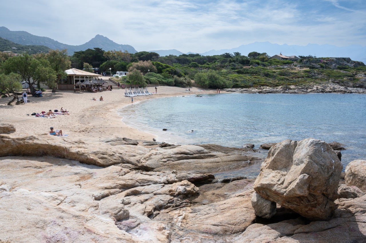 Blick auf eine größere Badebucht, die von einer grünen Landschaft umgeben ist, die in die Berge übergeht. Am Strand sind nur wenige Menschen und ein kleines Haus zu sehen.