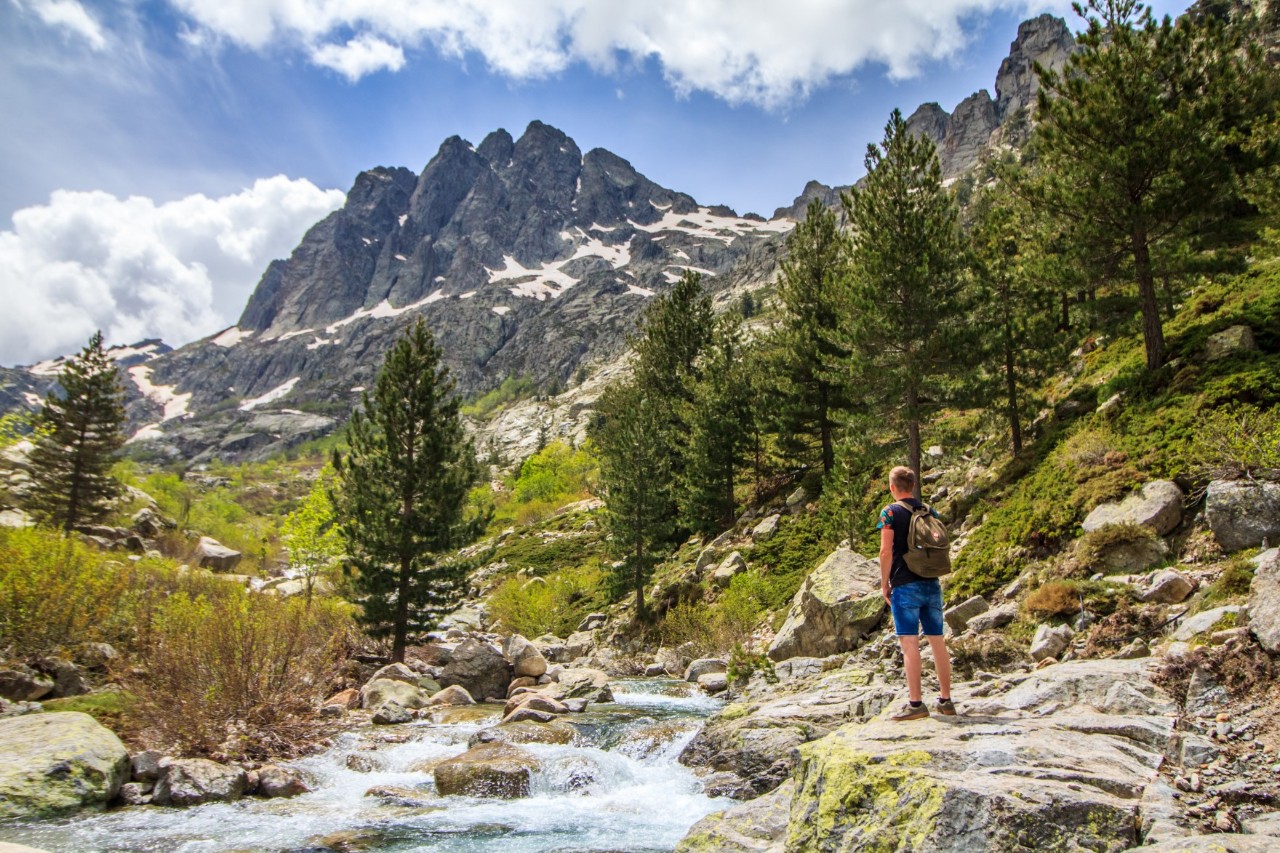 Blick auf einen hohen, leicht schneebedeckten Berg. Vor dem Berg verläuft ein Tal mit einem Fluss, Steinen und Bäumen. An der rechten Seite des Flusses steht ein Wanderer mit Rucksack.