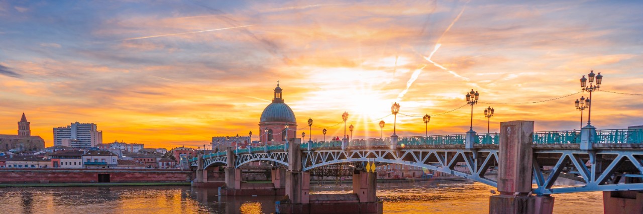 Sonnenuntergang mit Blick auf eine größere Brücke mit Laternen, die über einen Fluss verläuft. Auf der anderen Flussseite sind einige Häuser und die Kuppel einer Kirche zu sehen.