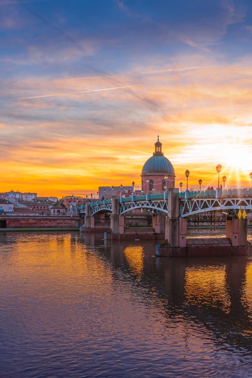 Sonnenuntergang mit Blick auf eine größere Brücke mit Laternen, die über einen Fluss verläuft. Auf der anderen Flussseite sind einige Häuser und die Kuppel einer Kirche zu sehen.