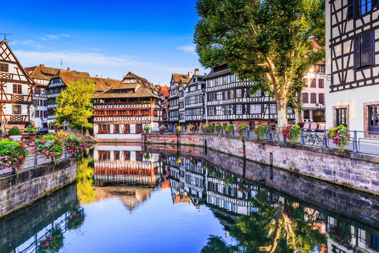 Traditionelle Fachwerkhäuser entlang eines ruhigen Kanals. Die Gebäude spiegeln sich in dem klaren Wasser des Kanals wider. Blumenampeln schmücken die Brücke und das Ufer, während ein großer Baum an der rechten Kanalseite die Szene belebt. Die Farben sind kräftig, der Himmel ist strahlend blau.