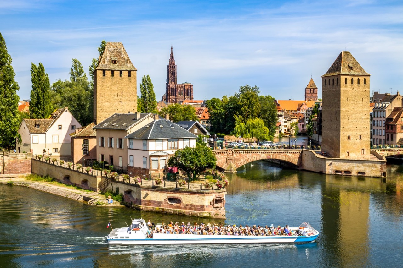 Das Bild zeigt eine Flusslandschaft mit einem belebten Ausflugsboot, das durch ruhiges Wasser fährt. Hinter dem Boot befinden sich zwei mittelalterliche Wachtürme sowie eine Brücke mit zwei Bögen, die den Fluss überquert. Im Hintergrund ist eine Altstadt mit traditionellen Gebäuden sowie das markante Straßburger Münster sichtbar. Der Himmel ist blau.