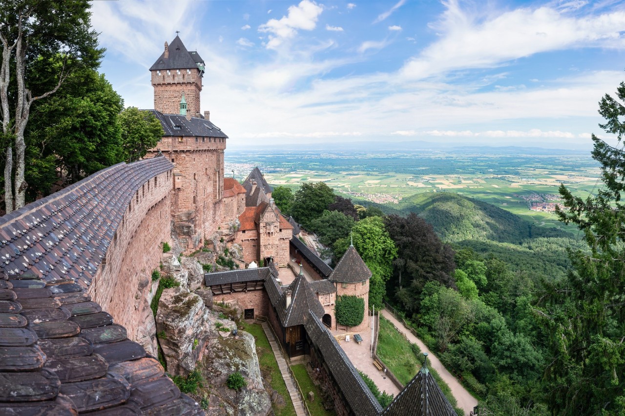 Auf dem Bild ist eine rötlich-sandfarbene Burg mit dunklen Dächern zu sehen, die hoch auf einem Hügel thront. Die Burgmauern und Türme erstrecken sich entlang eines steilen Abhangs. Sie ist von einer grünen Landschaft und bewaldeten Hängen bis zum Horizont umgeben. Der Himmel ist klar und blau.