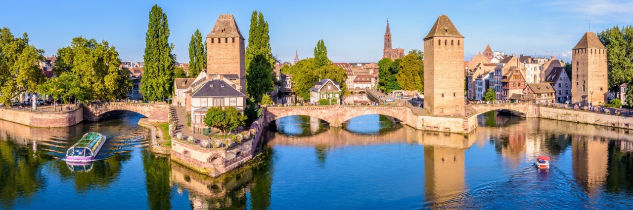 Das Bild zeigt eine Flusslandschaft mit drei markanten Wehrtürmen, zwei Booten auf dem Fluss, alten Brücken und historischen Gebäuden. Im Hintergrund erhebt sich neben vielen Häusern ein hoher Kirchenturm.