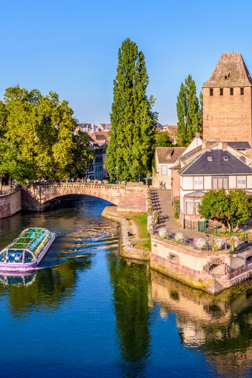 Das Bild zeigt eine Flusslandschaft mit drei markanten Wehrtürmen, zwei Booten auf dem Fluss, alten Brücken und historischen Gebäuden. Im Hintergrund erhebt sich neben vielen Häusern ein hoher Kirchenturm.