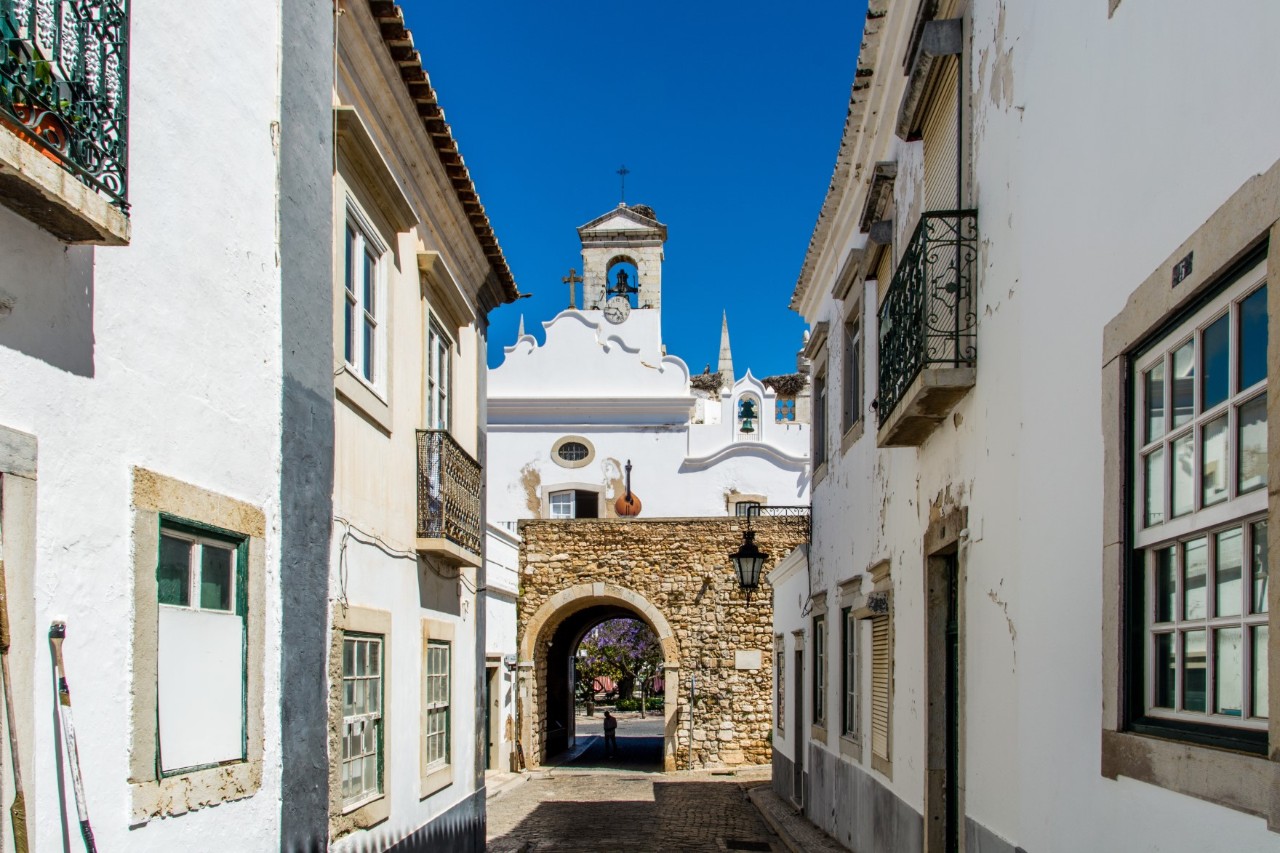 Weiße Häuser in der Altstadt von Faro, enge Häusergasse zur Steinmauer mit Durchgang, dahinter weiße Kirche mit Glockenturm. 