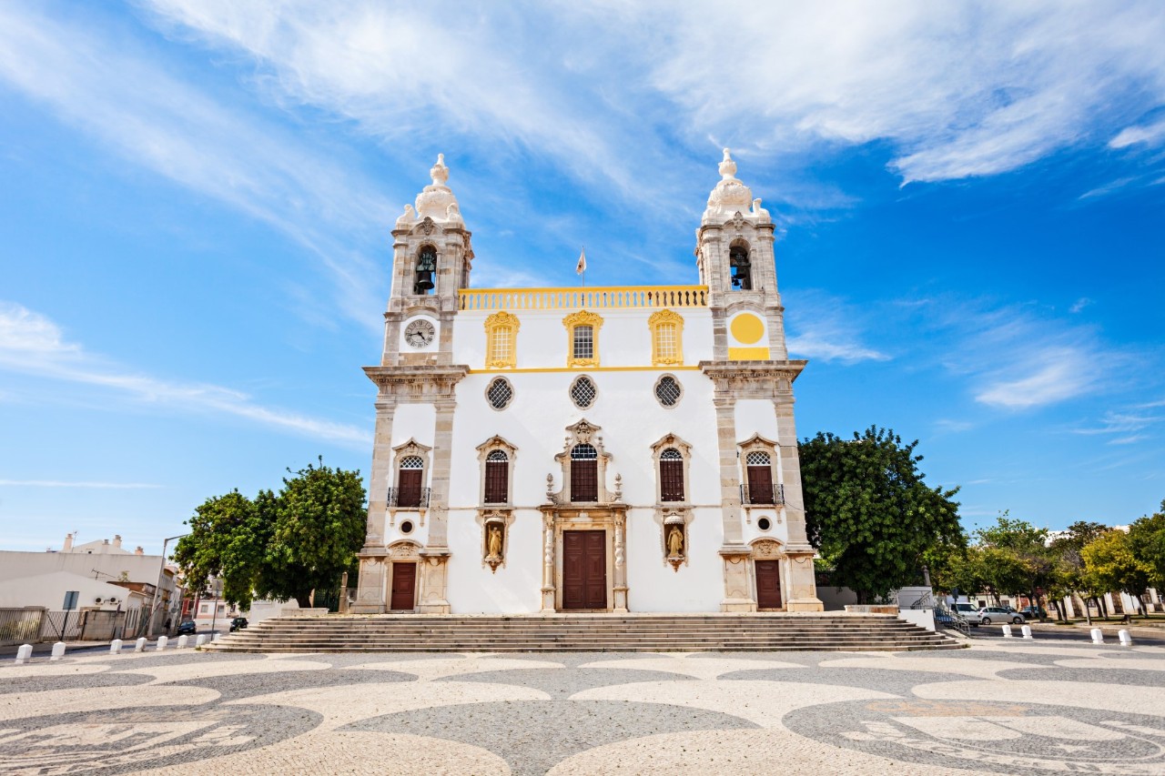 Kirche Carmo mit zwei Glockentürmen, goldenen Akzenten auf einem ruhigen Vorplatz, Bäume im Hintergrund. 