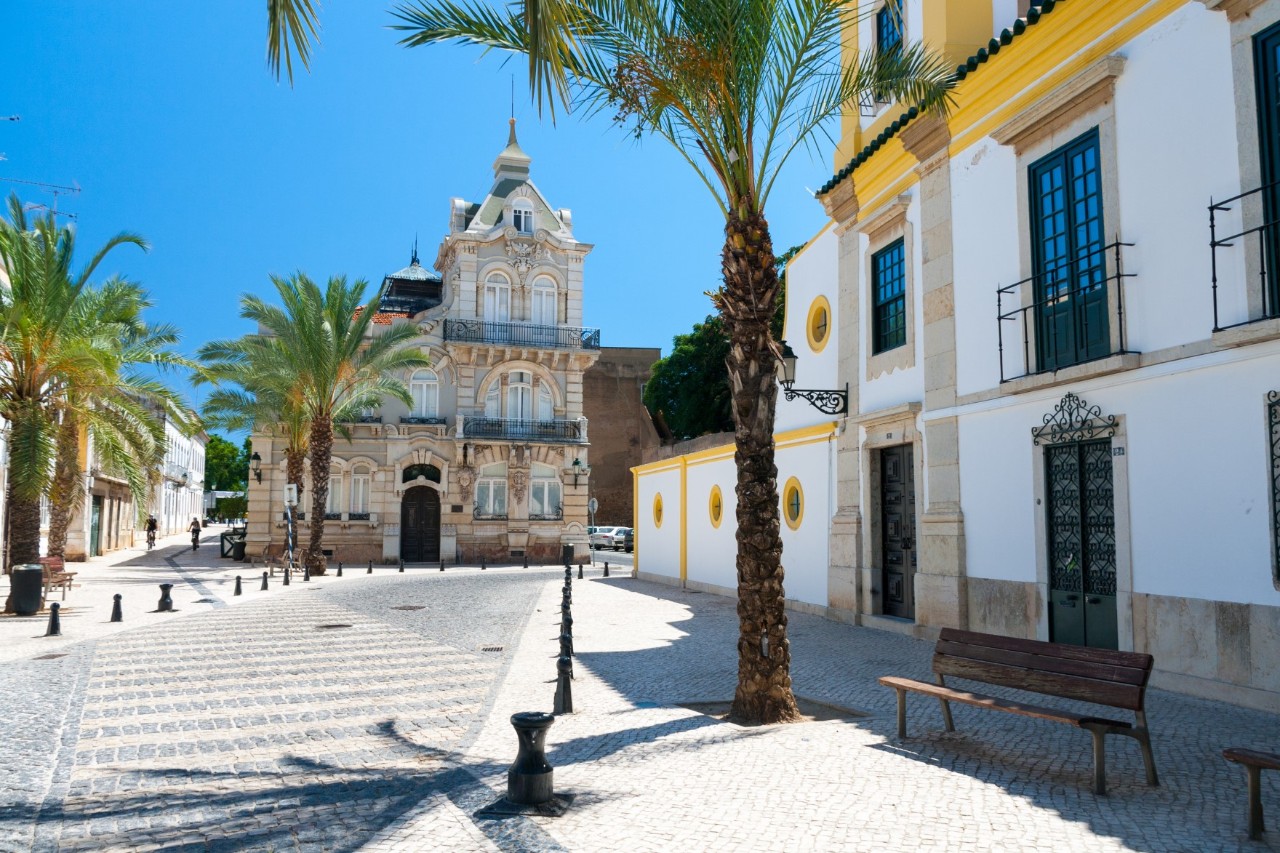 Unbelebter Platz in Faro mit Kirche im Hintergrund, Häusern im Stil von von Art Déco und South Modern, Palmen und Bank zum Ausruhen. 