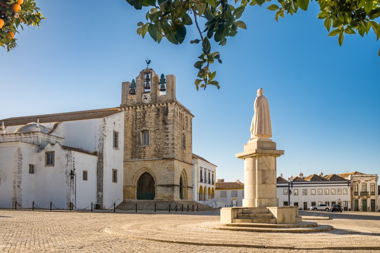 Kathedrale mit Glockenturm aus Stein und weiße Statue von Bischof D. Francisco Gomes de Avelar im Vordergrund, leerer Platz, weiße Häuser im Hintergrund. 
