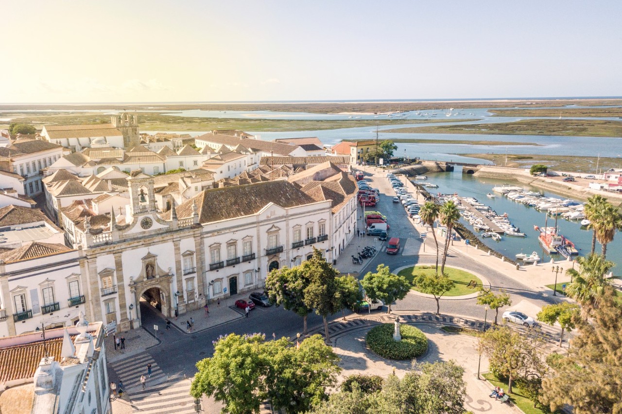 Weiße Häuser von Faros Innenstadt, kleiner bepflanzter Platz am Hafen, kleine Boote, in der Ferne Lagunen- und Seenlandschaft Naturschutzgebiet Ria Formosa. 