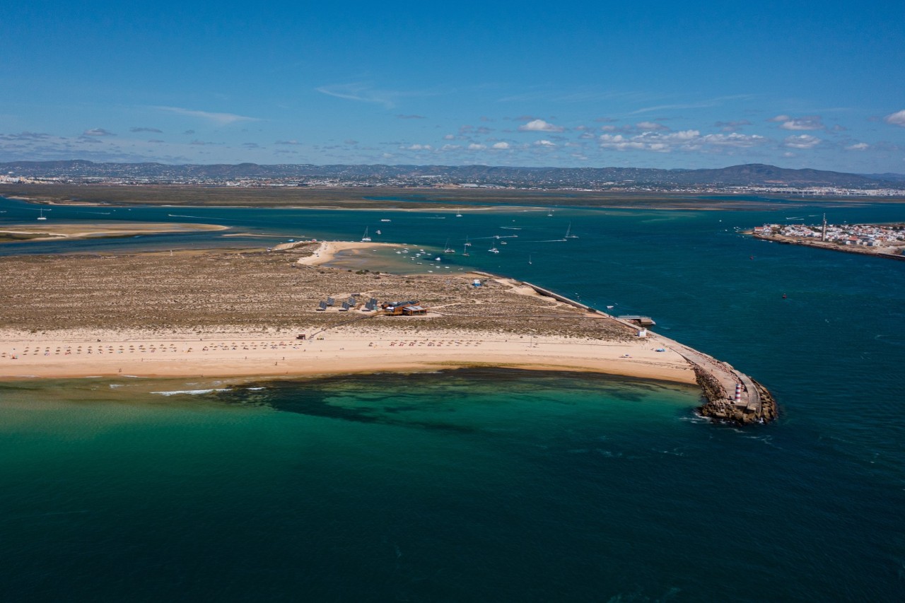 Vogelperspektive auf das Naturschutzgebiet Ria Formosa mit Flüssen und Sandstränden und -bänken, karge besiedelte Landschaft im Hintergrund. 