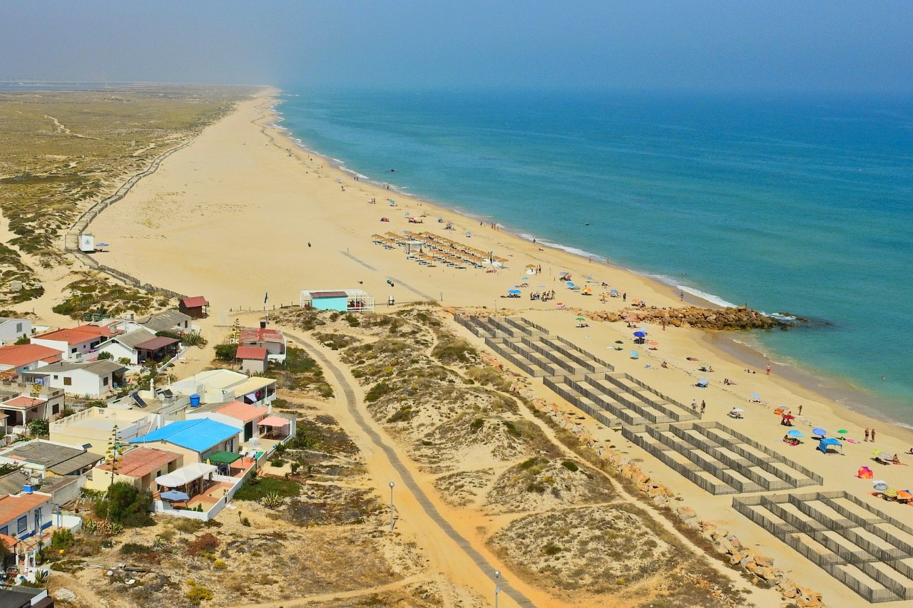 Sandstrand auf der Insel Ilha do Farol mit Ozean, Sonnenliegen und -schirme, kleine Häuser, karge Steppenlandschaft. 