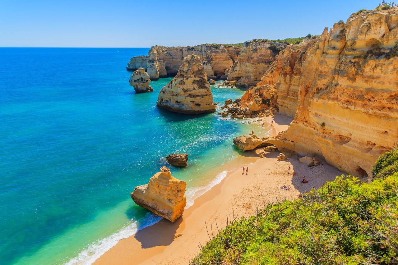 Sandstrand Marinha mit Steilküste und türkisblauem Wasser, einzelne Felsformationen, wenige Menschen am Strand.