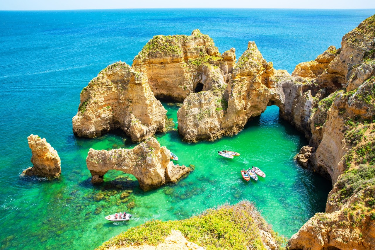 Felsformationen im Wasser an der Küste der Ponta da Piedade, türkisblaues Wasser, kleine Boote im Meer, spärlich bewachsene Steilküste. 