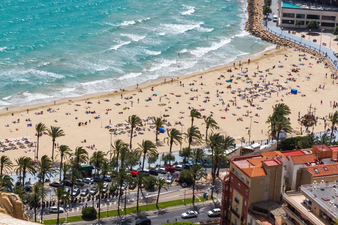 Blick von oben auf einen Sandstrand, der gesäumt ist von Palmen. Das azurblaue Meer schlägt leichte Wellen. Der Strand ist von zahlreichen Badegästen gut besucht. Unterhalb der Palmen sind parkende Autos und eine Straße sowie am rechten Bildrand Häuser zu sehen.