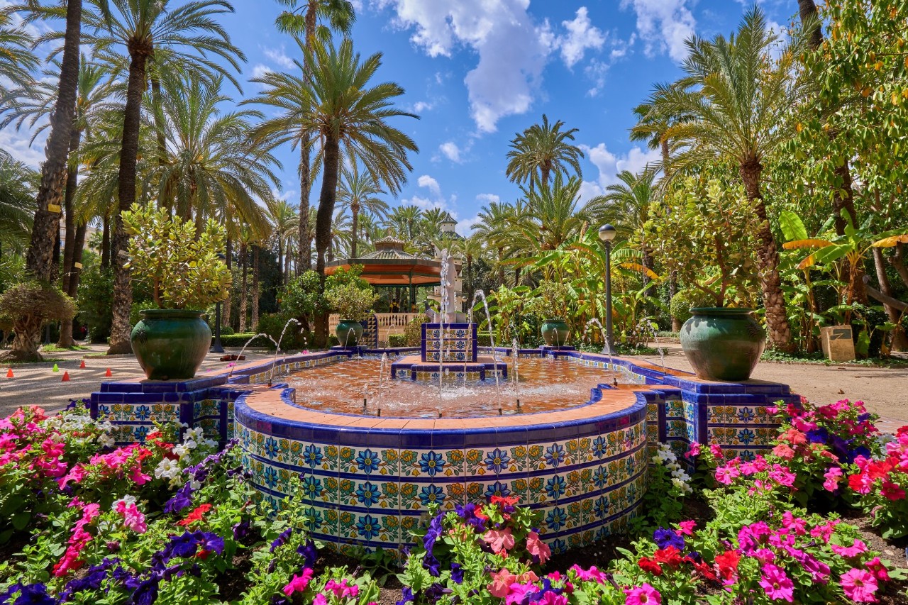 Ansicht eines Springbrunnens, der mit blauen und bunten Mosaikkacheln verziert und am Boden von bunten Blumen umrankt ist. Im Hintergrund sind verschiedene Palmenarten zu sehen.