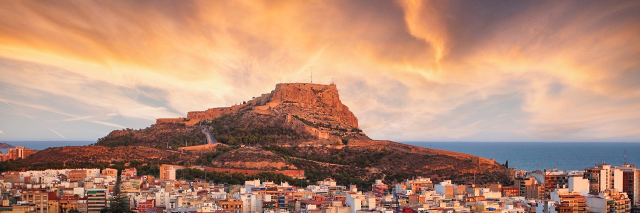 Luftbild von Alicante im Sonnenuntergang. Im Vordergrund ist das vom Sonnenuntergang rötlich erleuchtete Häusermeer zu sehen, im Hintergrund ein Berg mit einer Festung auf dem Gipfel, der ebenfalls rot strahlt.