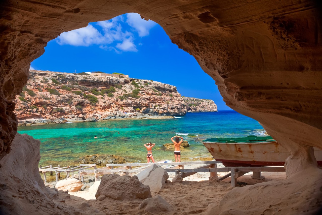 Das Bild zeigt den Blick aus einer Höhle auf eine Küstenlandschaft. Im Vordergrund stehen zwei Personen am Strand, die auf das Meer hinausschauen. Rechts ist ein altes Boot und im Hintergrund eine felsige Klippe zu sehen. 