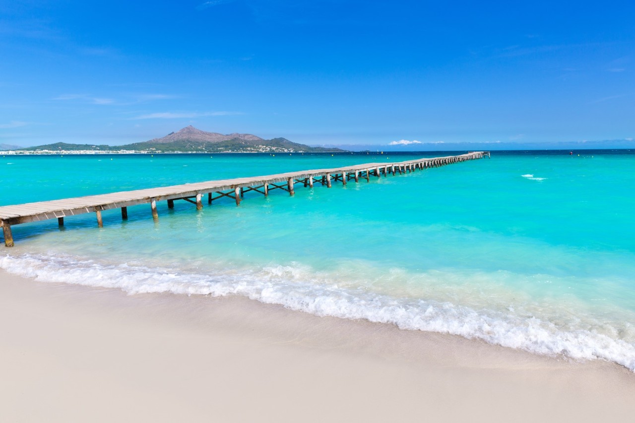 Türkisblaues Wasser und weißer Sandstrand mit einem langen Steg, der ins Wasser führt und einer Bergansicht im Hintergrund 