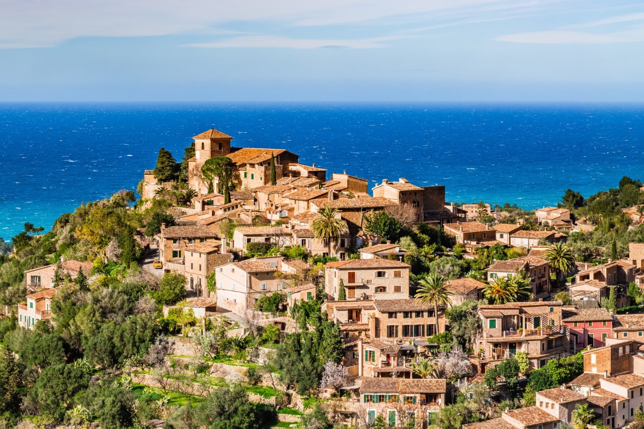 Luftbild des mallorquinischen Künstlerdorfs Deià, das an einen Berg gebaut ist, mit Meer im Hintergrund 