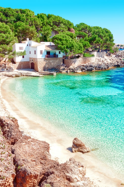 Türkisblaues Wasser und weißer Sandstrand auf Mallorca, eingerahmt von Bäumen und einem weißen Haus © pixelliebe/stock.adobe.com