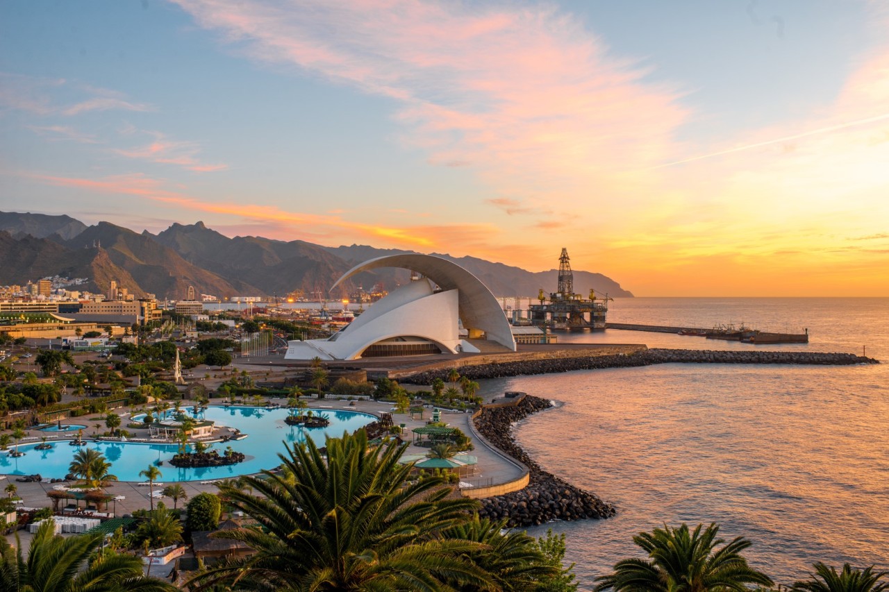 Santa Cruz bei Sonnenuntergang, geschwungenes Gebäude, Palmen, Meer, bergige Landschaft im Hintergrund 