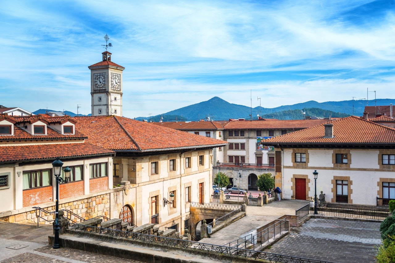Das Bild zeigt einen ruhigen Platz mit traditionellen Gebäuden und roten Ziegeldächern. Im Hintergrund ragt ein Uhrenturm empor. Dahinter sind bewaldete Hügel und Berge zu sehen. 