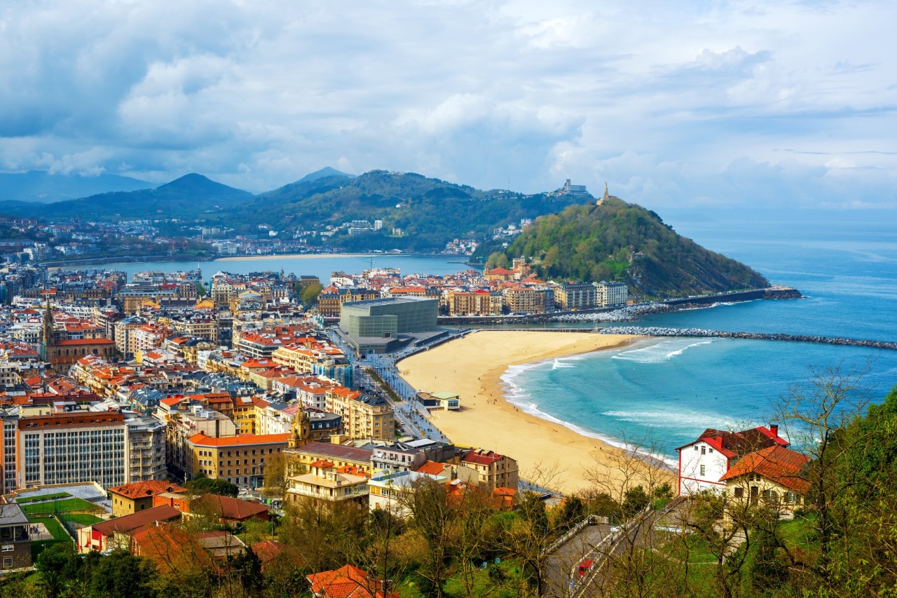 Das Bild zeigt eine Panoramaansicht einer Küstenstadt mit vielen Häusern und einem breiten Sandstrand. Im Hintergrund erstrecken sich grüne Hügel. 