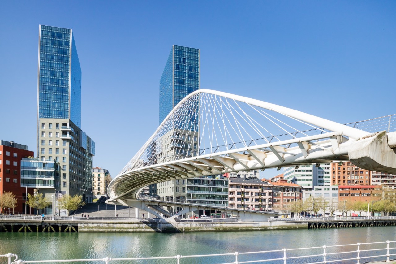 Das Bild zeigt eine weiße, bogenförmige Fußgängerbrücke, die über einen Fluss führt. Im Hintergrund erheben sich zwei markante Wolkenkratzer, eingebettet in eine städtische Kulisse.