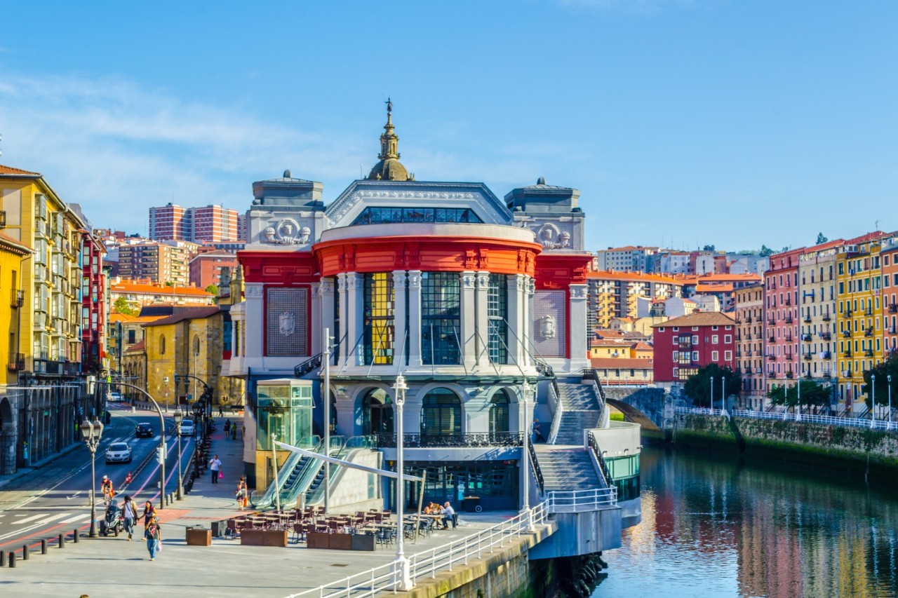 Das Bild zeigt ein Gebäude im Art-Déco-Stil mit auffälligen roten und weißen Elementen sowie großen Fenstern. Im Vordergrund ist das Flussufer zu sehen, im Hintergrund befinden sich bunte Häuser der Altstadt.