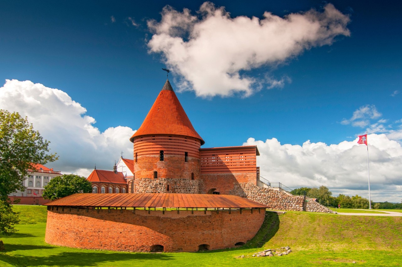 Rote Burg mit rundem Turm und Mauer, umgeben von einer Wiese 