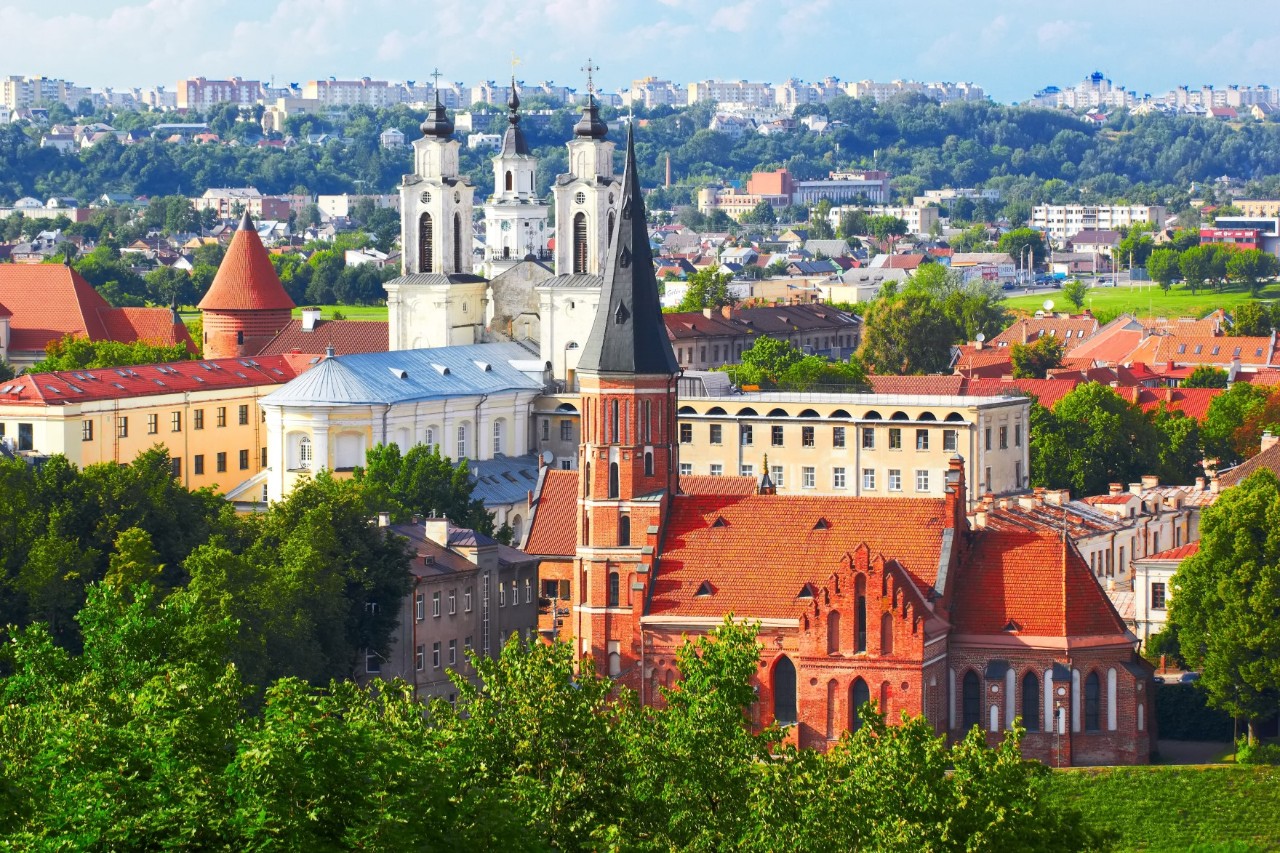 Blick über eine Stadt mit Bäumen, einer Kirche und großen Häusern im Vordergrund und einer weiteren Stadtansicht im Hintergrund 