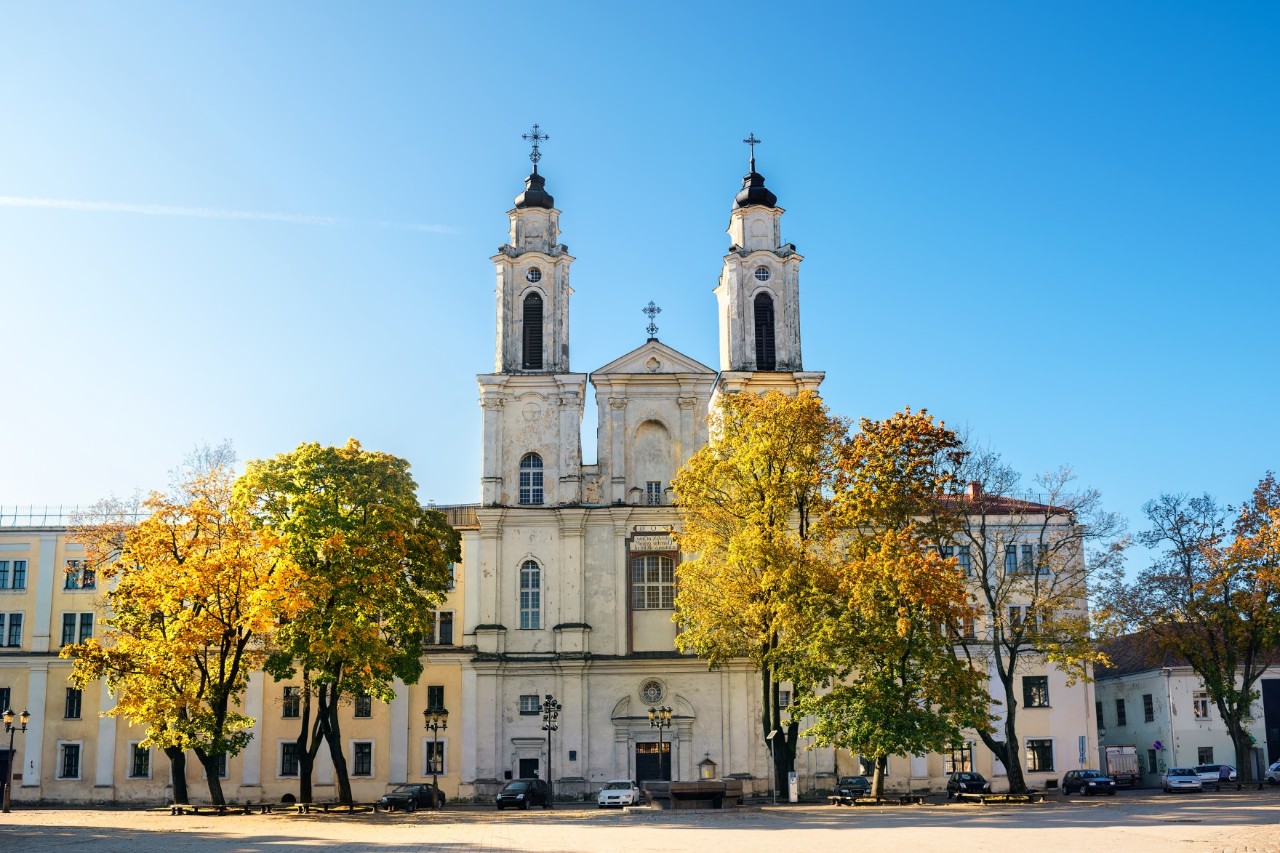 Weiße Kirche mit zwei Türmen und herbstlich gefärbten Bäumen vor dem Gebäude 