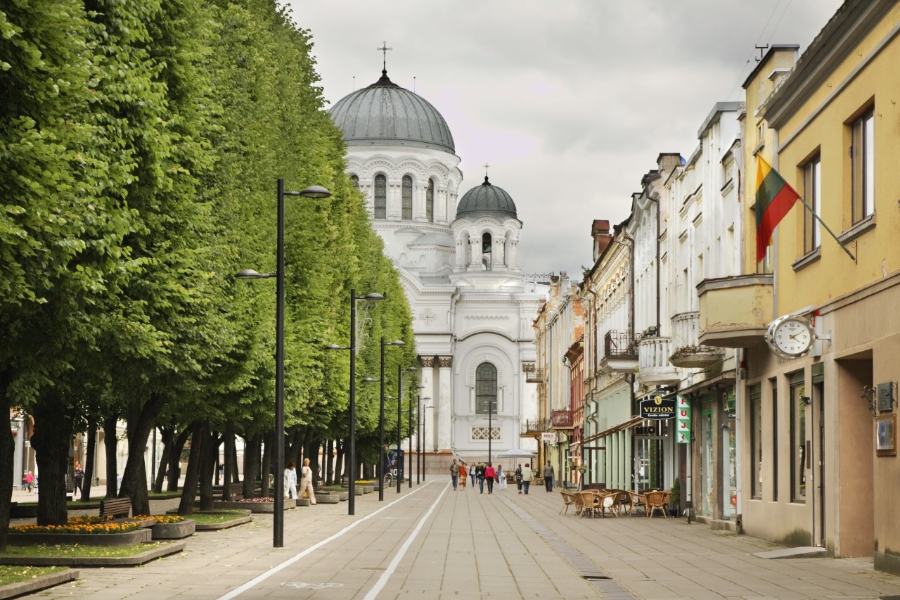 Fußgängerzone mit Häusern rechts im Bild und Bäumen links, einigen Menschen und weißer Kirche mit zwei Türmen und runden Kuppeln am Ende der Straße 