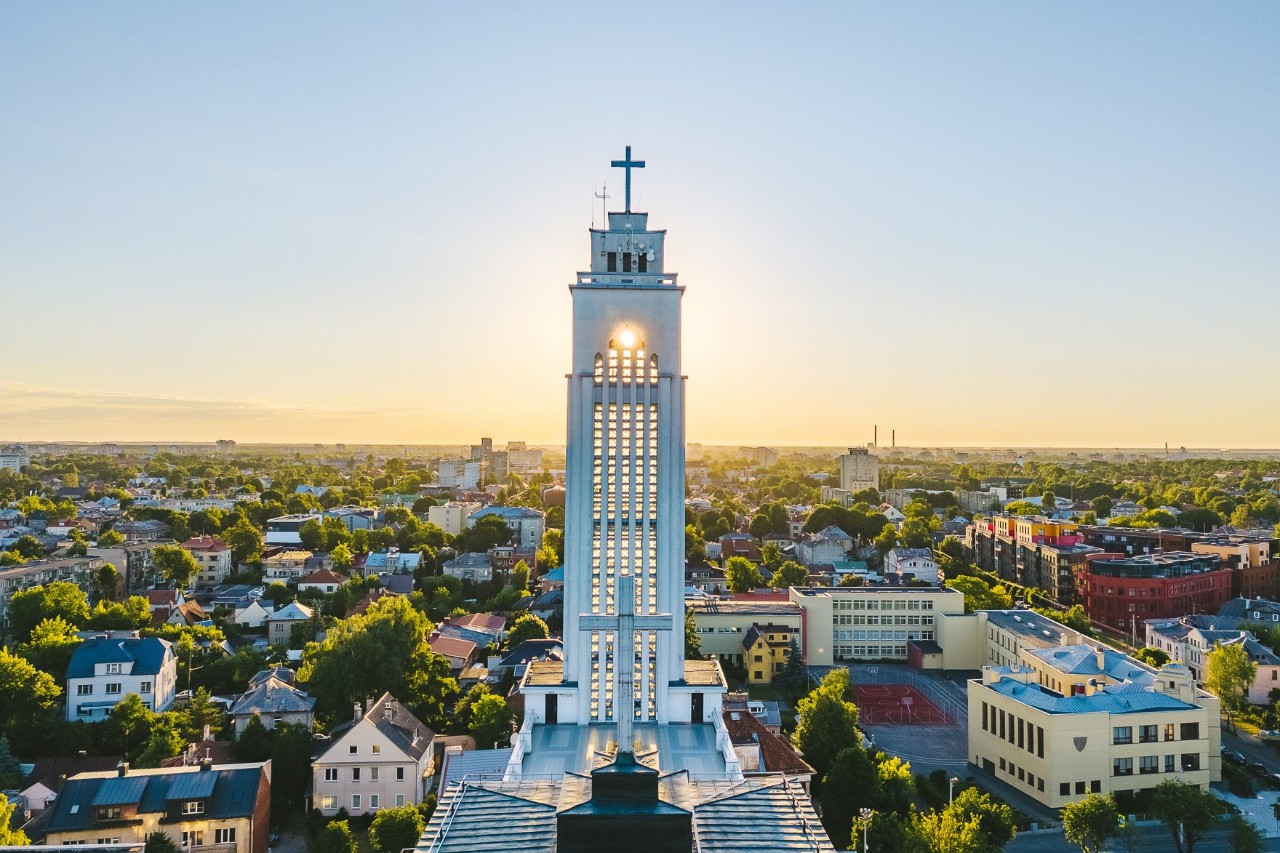 Luftbild mit Blick auf eine Stadt mit Horizontlinie bei Sonnenaufgang. Markant im Bild ist ein weißer, sehr hoher Kirchturm inmitten von Häusern und Bäumen 