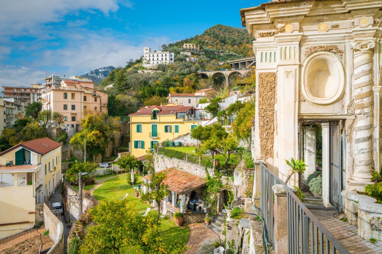 Ein Garten zwischen mehreren Wohnhäusern in einer Stadt, der sich terrassenartig auf zwei Ebenen erstreckt. Mehrere Bäume und Büsche sind zu sehen, außerdem eine überdachte Sitzgelegenheit mit davorstehenden Liegestühlen. Im Vordergrund ist ein Teil eines imposanten, antiken Gebäudes mit einer davor gebauten Terrasse mit Balkongeländer. Am Horizont erhebt sich ein spitz zulaufender Hügel, auf den mehrere Häuser gebaut sind.
