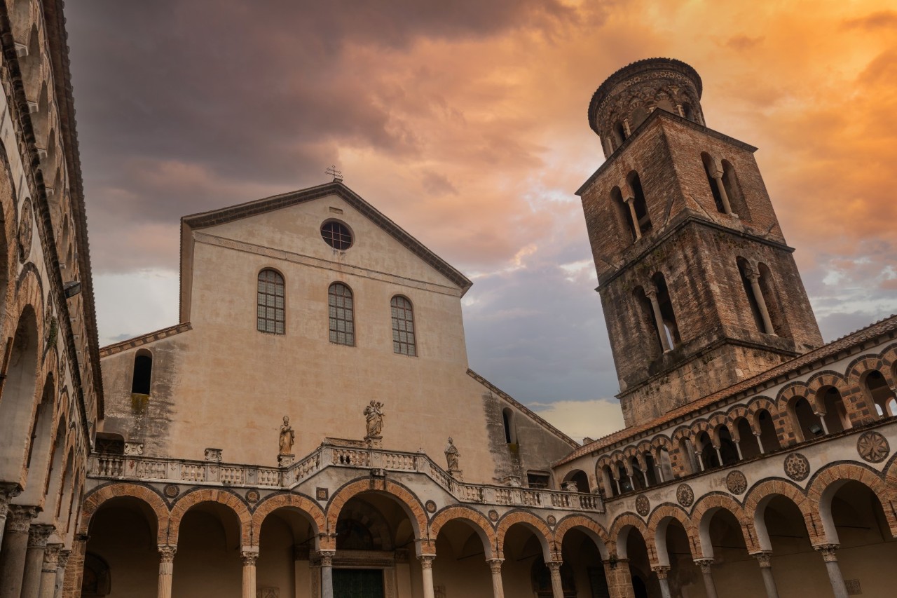 Bild aus dem Innenhof einer großen Kathedrale. Der Hof ist mit Arkaden gesäumt, im Bildzentrum ist das Hauptschiff der Kirche, unter dessen Satteldach sind drei Bogenfenster und unter der Spitze ein rundes Fenster zu sehen. Rechts erhebt sich ein viereckiger Turm mit Bogenfenstern und einem Rundbogenaufbau. Der Himmel leuchtet im Abendrot.