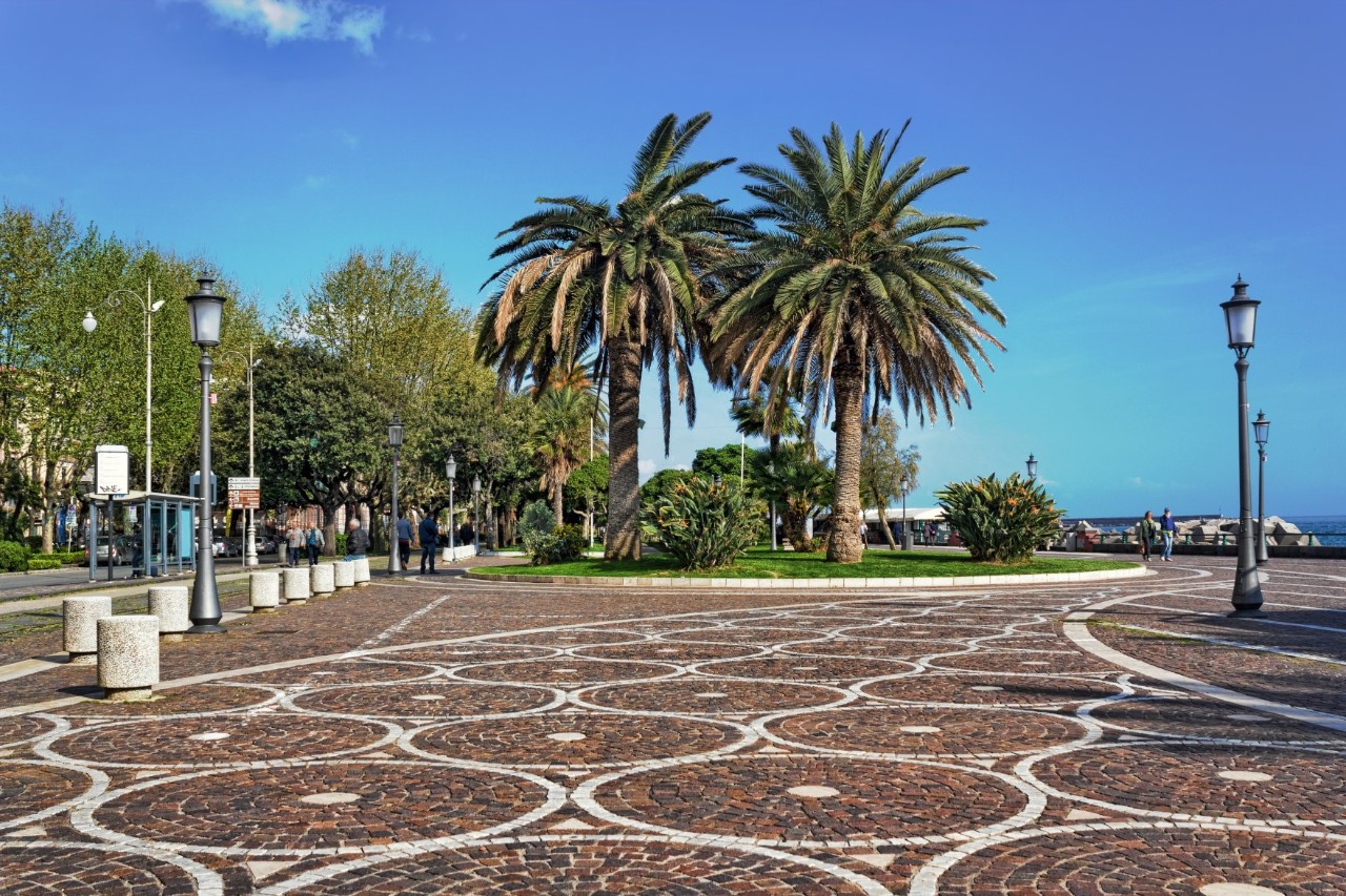 Platz mit einem terrakottafarben-weiß gemusterten Mosaikboden. Rechts und links stehen historische Straßenlaternen, in der Bildmitte ist ein grasbewachsenes Rondell mit mehreren Palmen zu sehen. Links im Bild sind Bäume, am rechten Bildhintergrund lässt sich das Meer erahnen.