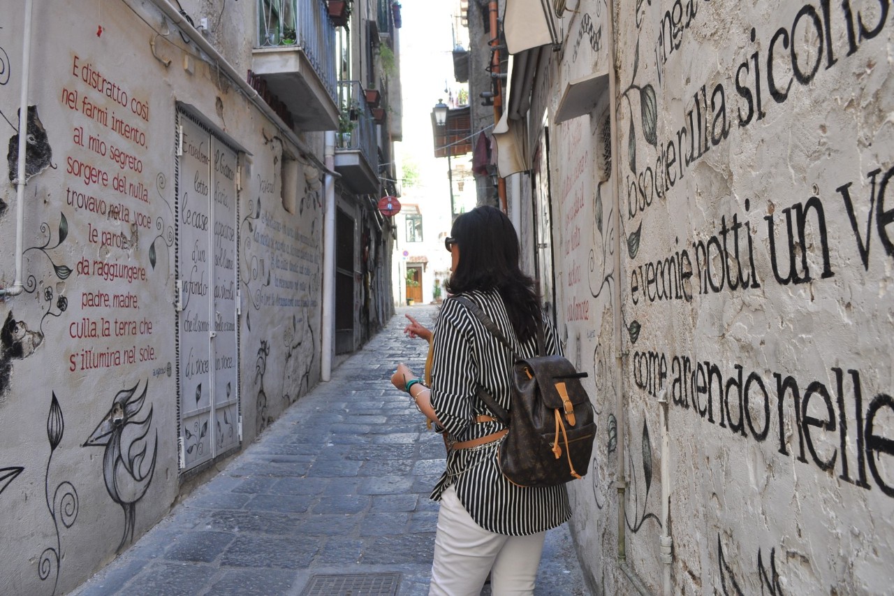 Blick in die gepflasterte, schmale Gasse einer Altstadt. Im Bildvordergrund steht eine Frau mit Rucksack, deren Rücken dem Bild zugewandt ist. Auf den Häuserwänden rechts und links der Gasse sind Gedichte und Zeichnungen zu sehen.