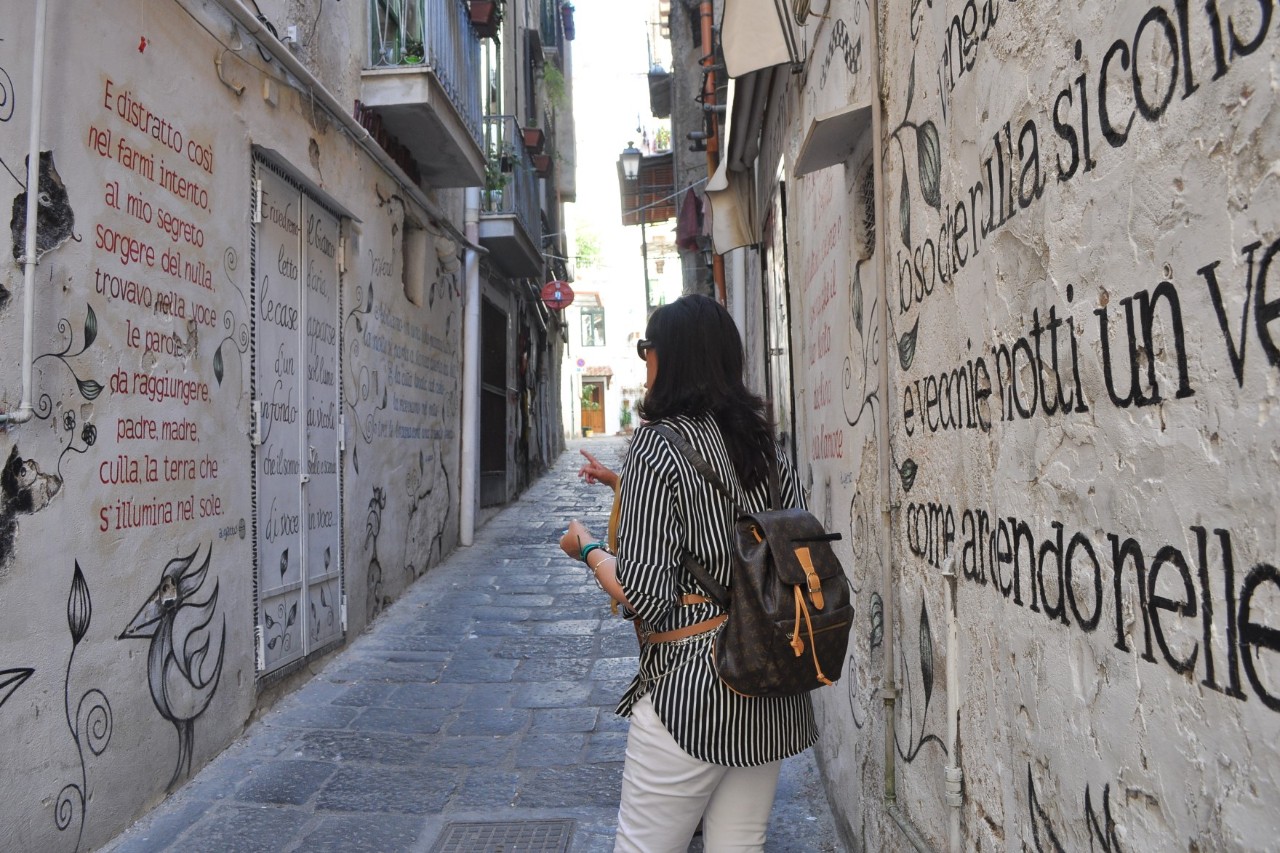 Gepflasterte, schmale Gasse einer Altstadt. Im Bildvordergrund steht eine Frau mit Rucksack, deren Rücken dem Bild zugewandt ist. Auf den Häuserwänden rechts und links der Gasse sind Gedichte und Zeichnungen zu sehen.
