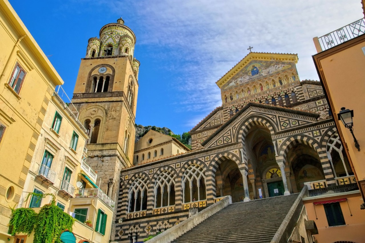 Eine breite Treppe führt nach oben zu einer prächtigen Kathedrale, deren Fassade durch Streifen und Mosaikmuster aus schwarzem und weißem Marmor geprägt ist und Bögen und Säulen hat, die an arabische Architektur erinnern. Links der Hauptfassade erhebt sich ein viereckiger Turm aus Sandstein, der rundum mit Bogenfenstern versehen ist. Links und rechts der Treppe sind gelbe Häuser sichtbar.