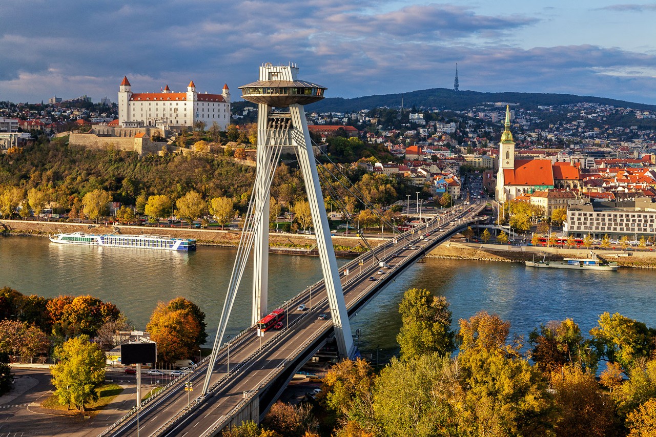 Eine asymmetrische Stahl-Schrägseilbrücke führt über die Donau. Über der Brücke ragt ein schräger Pylon mit einem UFO-förmigen Turmaufsatz. Im Hintergrund befindet sich die Altstadt, umgeben von herbstlich gefärbten Bäumen und dem dicht bebauten Stadtviertel. 