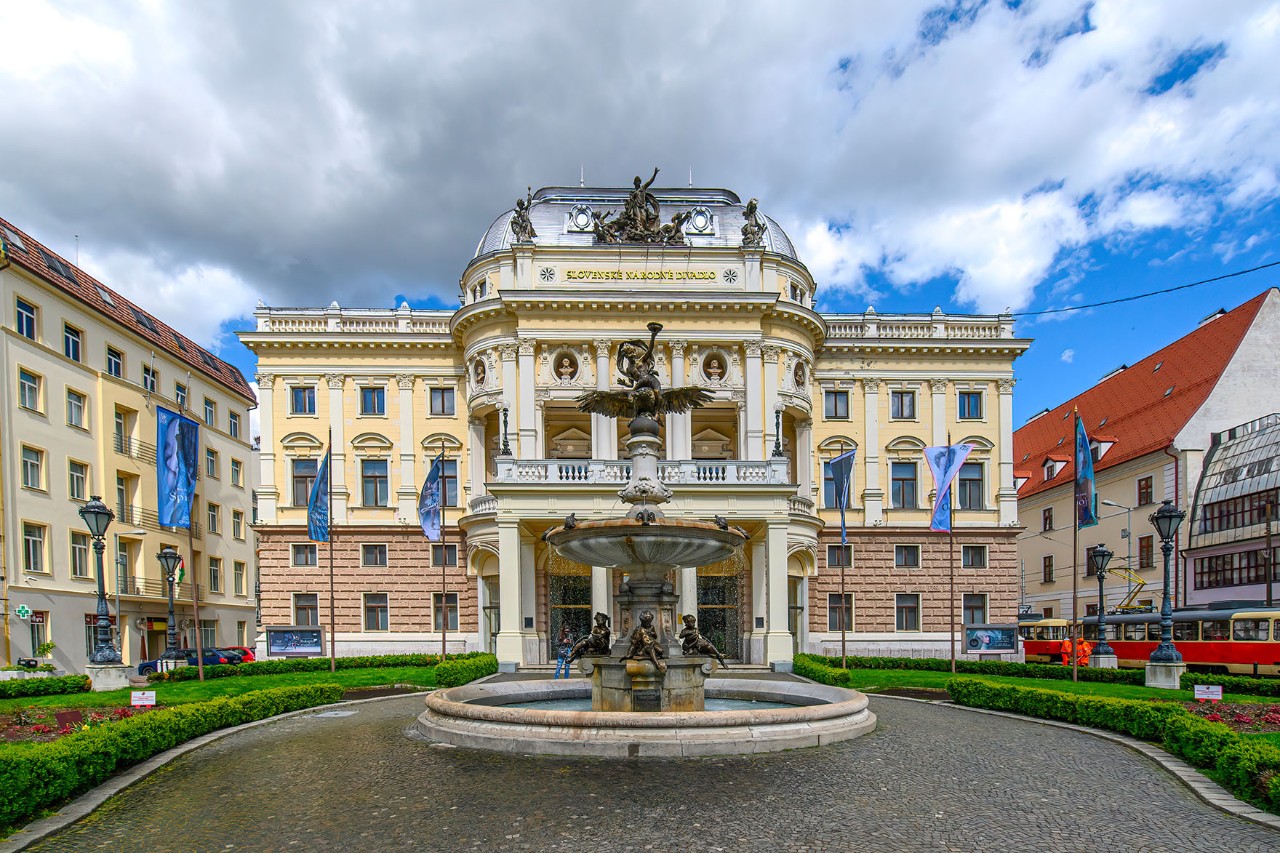 Statue auf einem Sockel mit Brunnen im Vordergrund, umgeben von einer grünen Parkanlage. Bäume und Wege rahmen den Platz bei sonnigem Wetter.