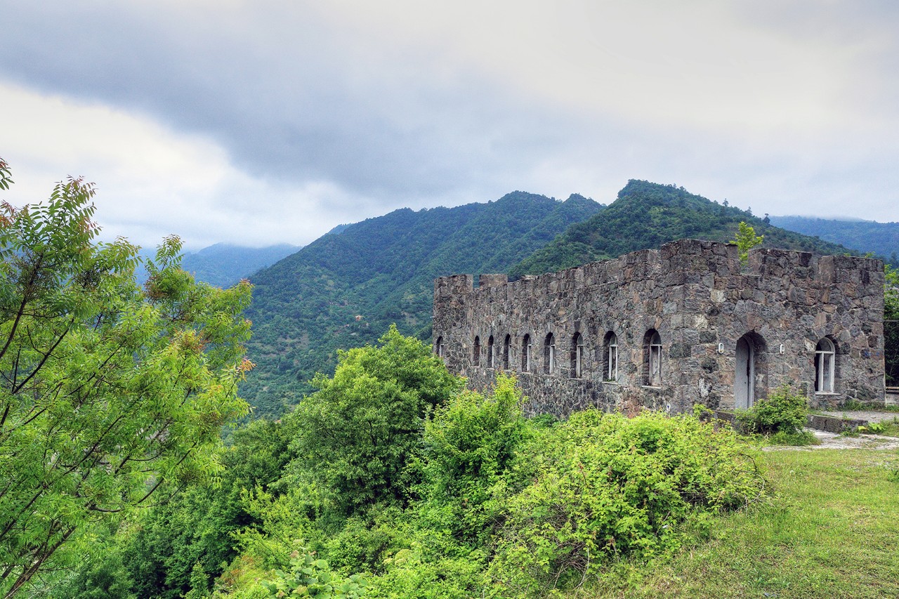 Das Bild zeigt die Ruine eines alten, rechteckigen Steinbaus mit Rundbogenfenstern inmitten grüner, bewaldeter Berge. 