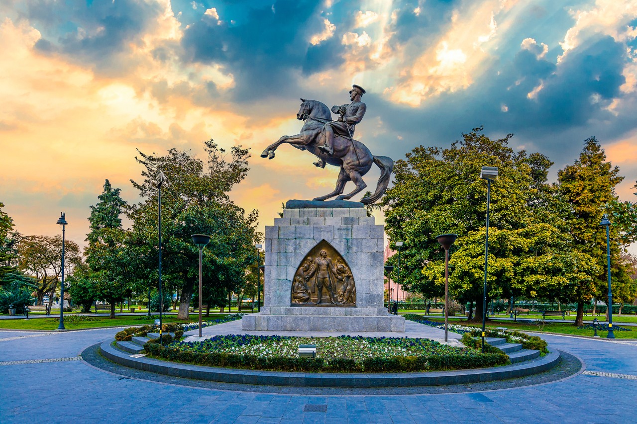 Reiterstatue auf einem steinernen Sockel mit goldenem Relief,  umgeben von Bäumen im Park. Bedeckter Himmel mit Sonne, die durch die Wolken scheint.