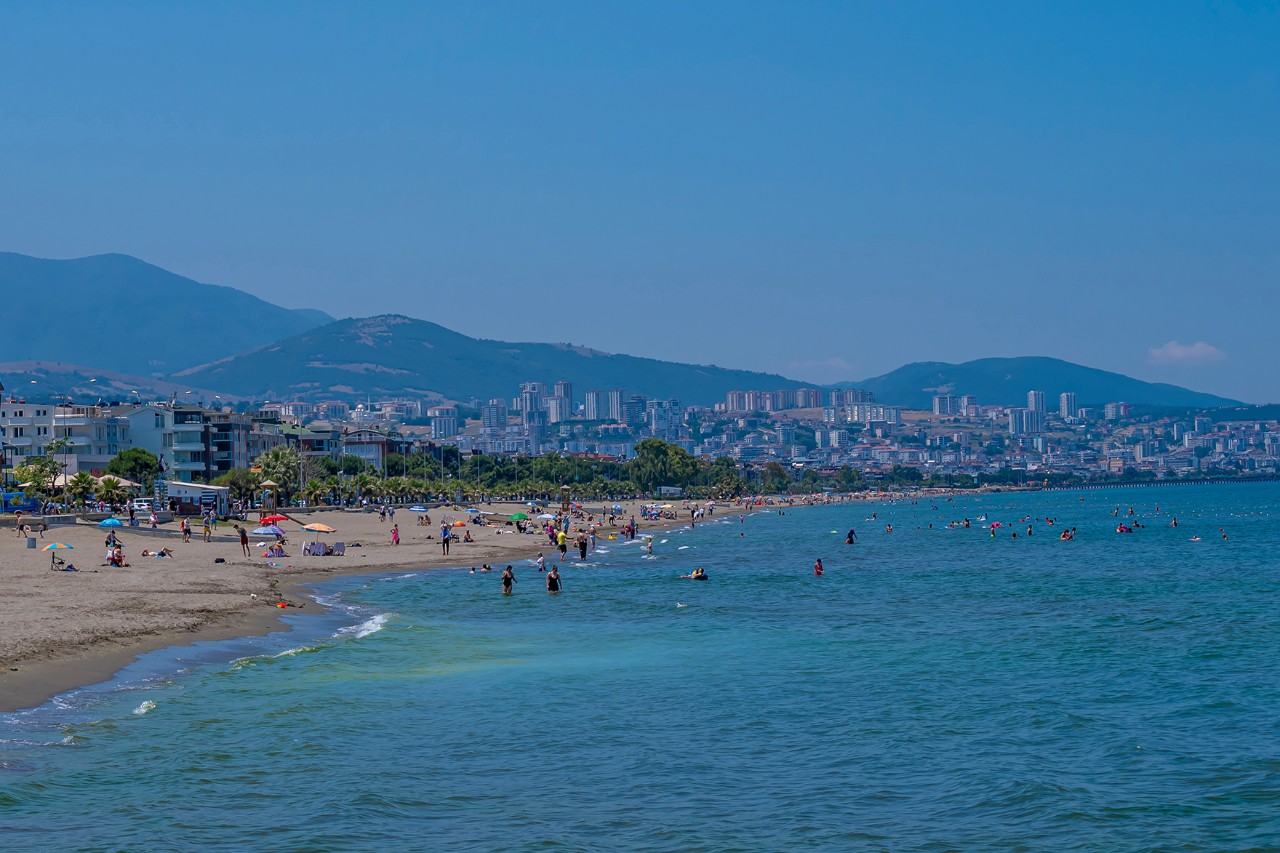 Belebter Strand mit Badegästen im Wasser und am Ufer. Im Hintergrund ist eine Stadt mit Hochhäusern zu sehen, eingebettet in eine bergige Landschaft. 
