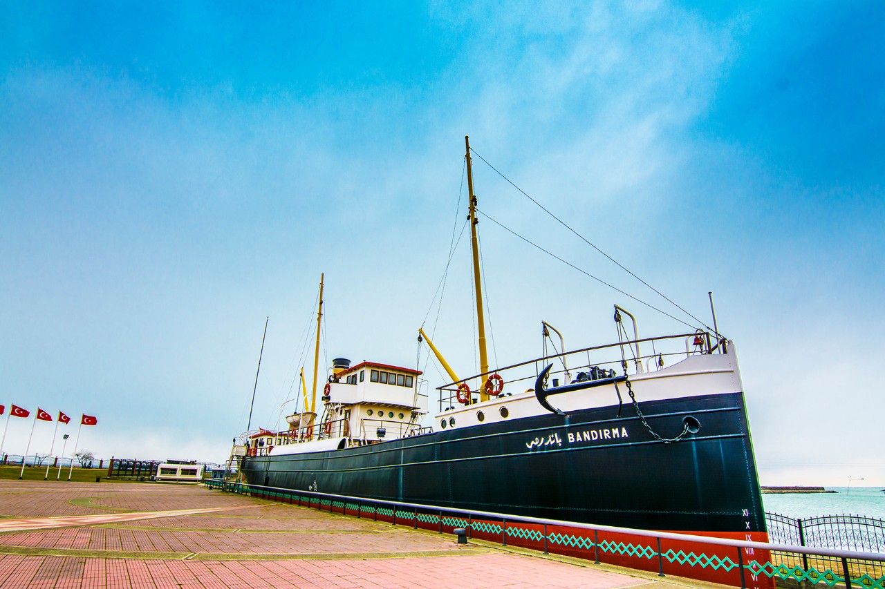 Ansicht eines alten Dampfschiffes, fest vertäut am Ufer bei klarem Himmel, mit türkischen Flaggen im Hintergrund und einem leeren, gepflasterten Platz im Vordergrund.