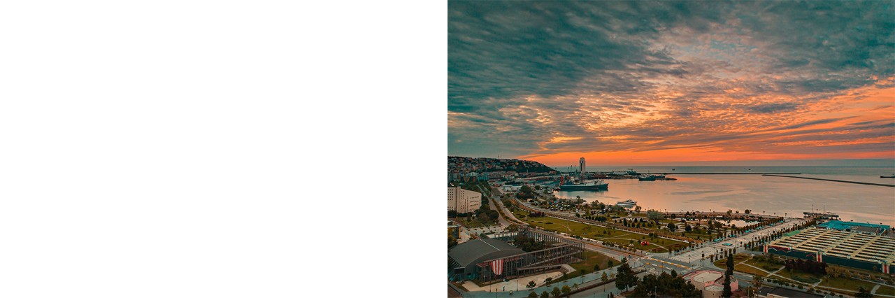 Aufnahme einer Küstenstadt bei Sonnenuntergang mit Park, Häusern, Straßen und einem Hafen mit Schiffen. Der Himmel ist mit orange- und blau gefärbten Wolken bedeckt. 