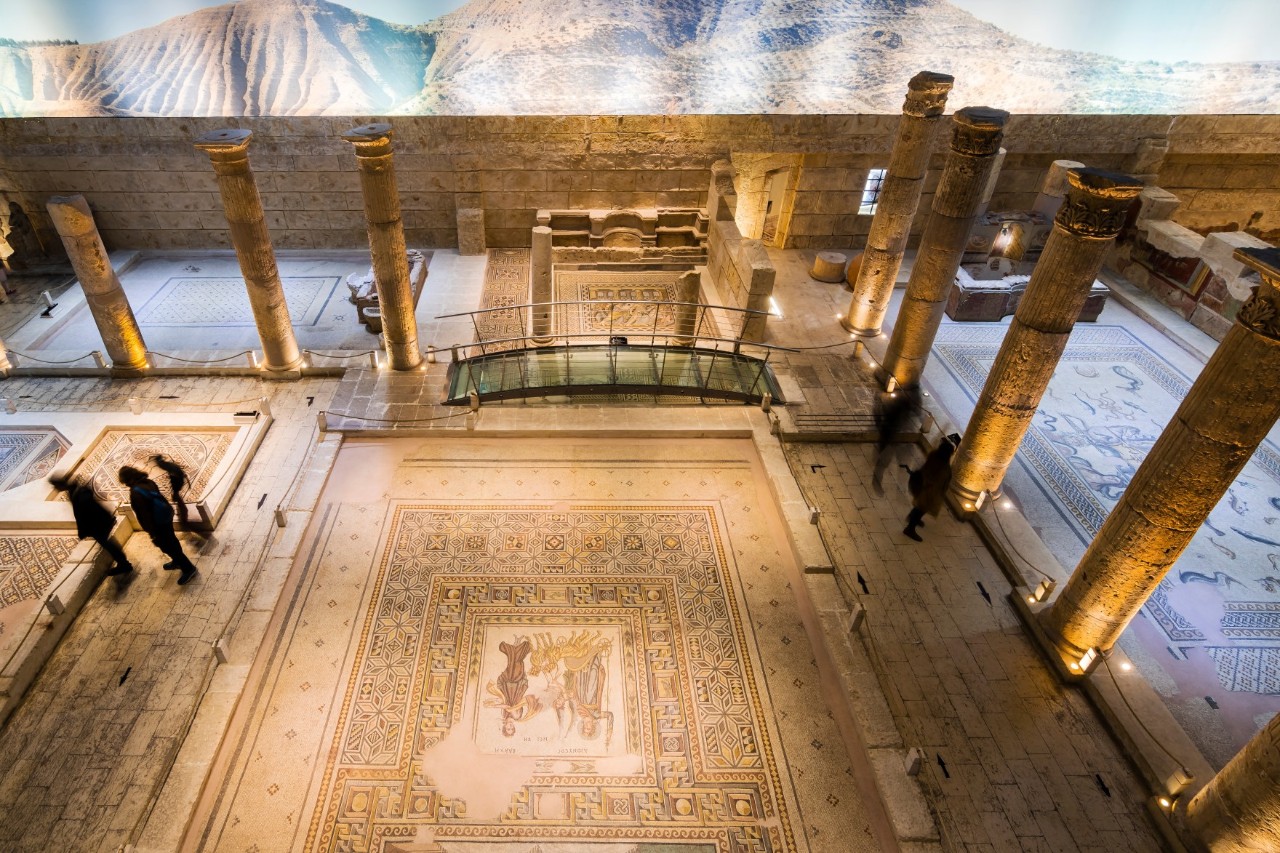 Innenaufnahme aus der Vogelperspektive eines Museums mit großflächigen Bodenmosaiken, antiken Säulen und gedämpfter Beleuchtung. Einige Menschen betrachten die Ausstellung.