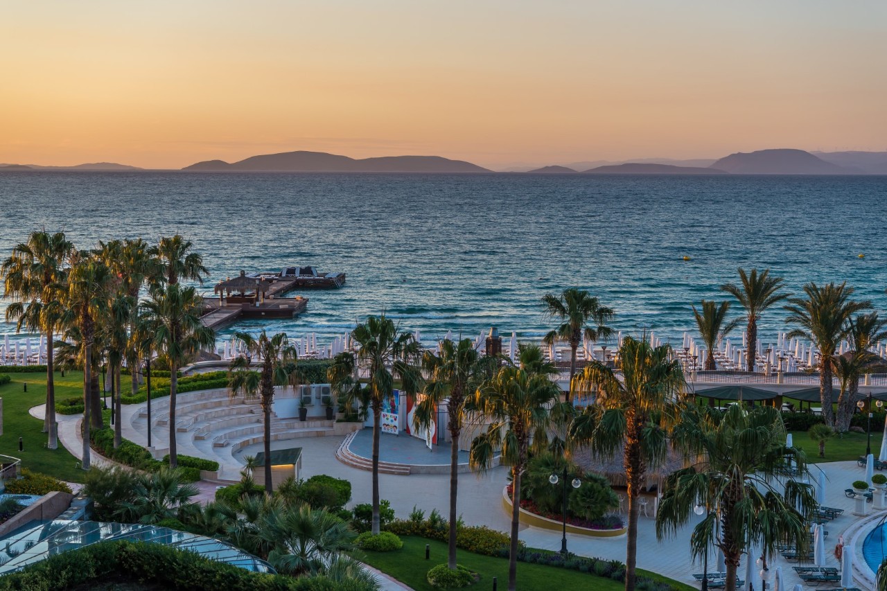 Blick auf eine am Meer gelegene Hotelanlage mit einer Bar, zahlreichen Palmen, Rasenfläche und einem Pool, dessen Rand rechts im Bild sichtbar ist. Auf der linken Bildseite ragt eine Steganlage ins Meer. Zusammengeklappte Sonnenschirme stehen am Strand. Am Horizont ist eine hügelige Landschaft sichtbar. Die Sonne ist schon untergegangen. Der Himmel ist noch orange.